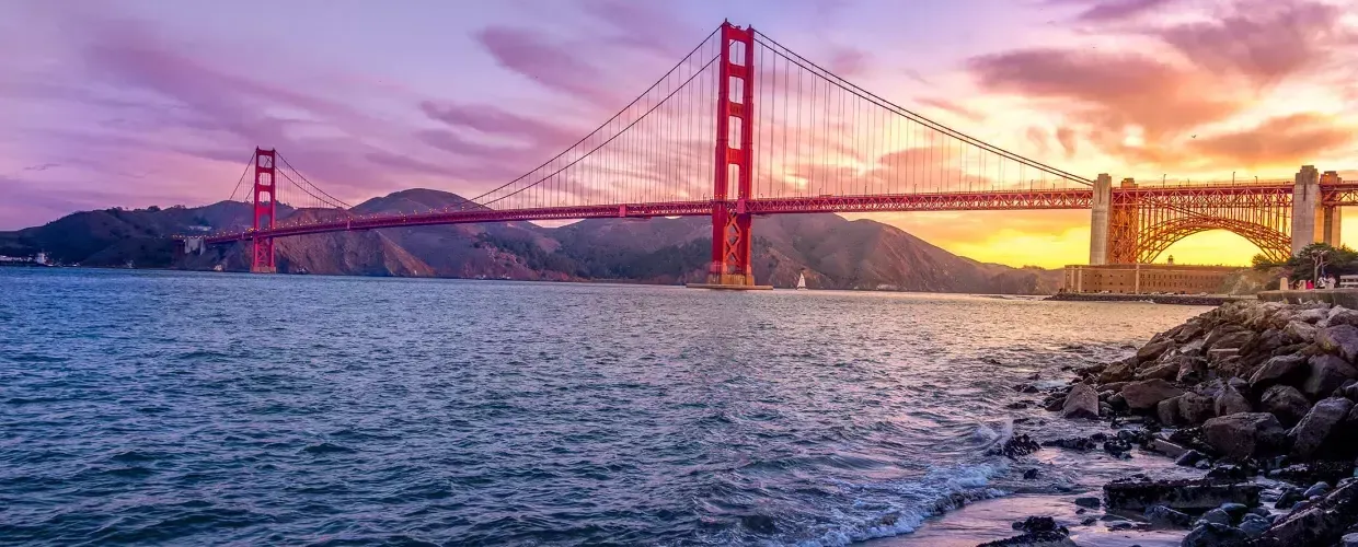 的 金门大桥 at sunset with a multicolored sky 和 San Francisco Bay in the foreground.