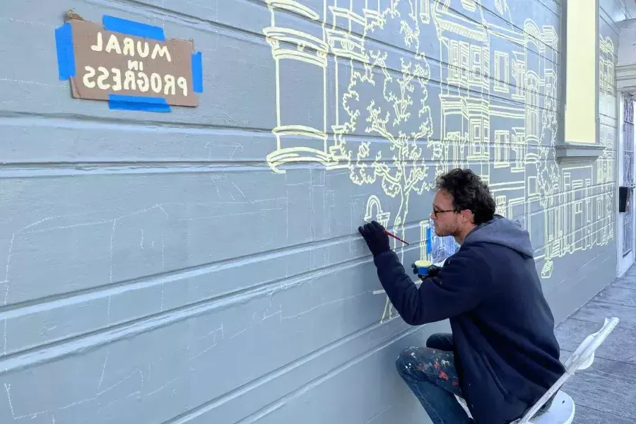 一位艺术家在教会区一栋建筑的侧面画了一幅壁画。, an dem ein Schild mit der Aufschrift „Mural in Progress“ angebracht ist. San Francisco, Kalifornien.